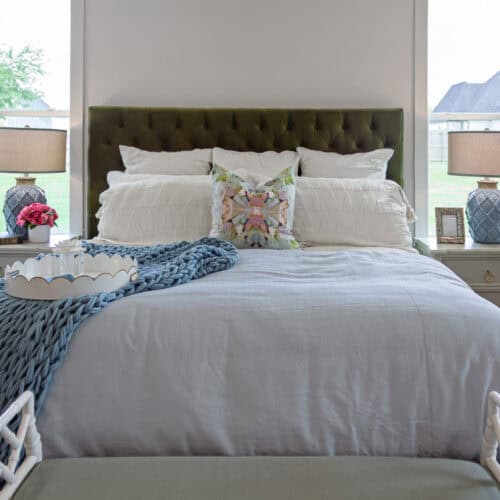 Luxurious Bedroom With White Bedding, Green Tufted Headboard, And Matching Nightstands Featuring Lamps, Decorative Pillows, And Fresh Flowers, Set In A Bright Room With Large Windows Overlooking A Backyard.