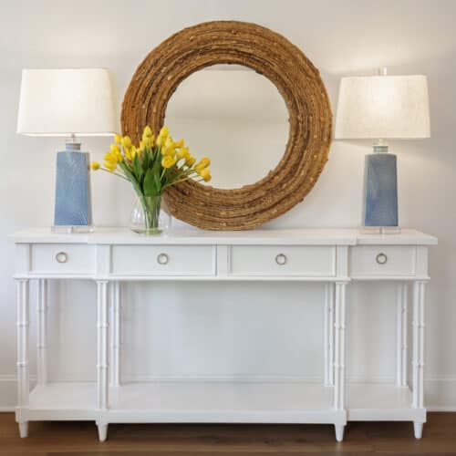 Elegant White Console Table With Drawer Storage, Adorned With A Glass Vase Of Yellow Tulips, Two Blue Table Lamps With Cream Shades, And A Large Rustic Wooden Mirror Above In A Contemporary Home Decor Setting.