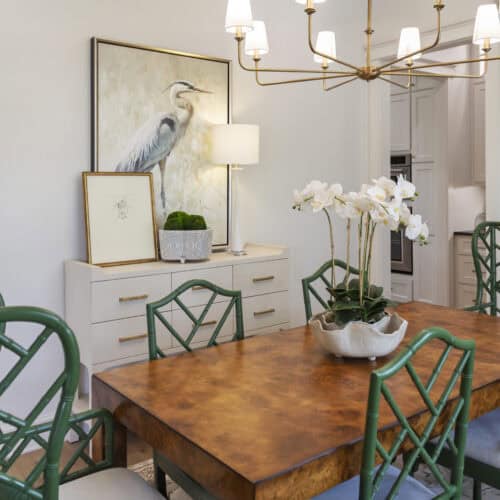 Elegant Dining Room With A Rustic Wooden Table, Green Bamboo-Style Chairs, White Orchid Centerpiece, And Stylish Decor Including A Bird Artwork And Modern Chandelier.
