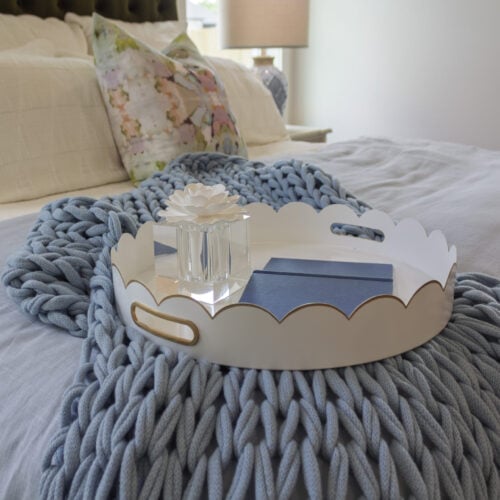 Decorative White Tray With Scalloped Edges On A Cozy Bed, Featuring A Glass Flower Sculpture And Two Navy Blue Notebooks, Accentuated By A Chunky Knit Blue Blanket.