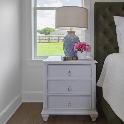 Elegant White Three-Drawer Nightstand With Gold Ring Pulls, Placed Beside A Bed With A Dark Green Tufted Headboard, Topped With A Decorative Blue Lamp, Pink Peony Flowers, And A Book, Set In A Bright Bedroom With A Window View Of A Green Backyard.