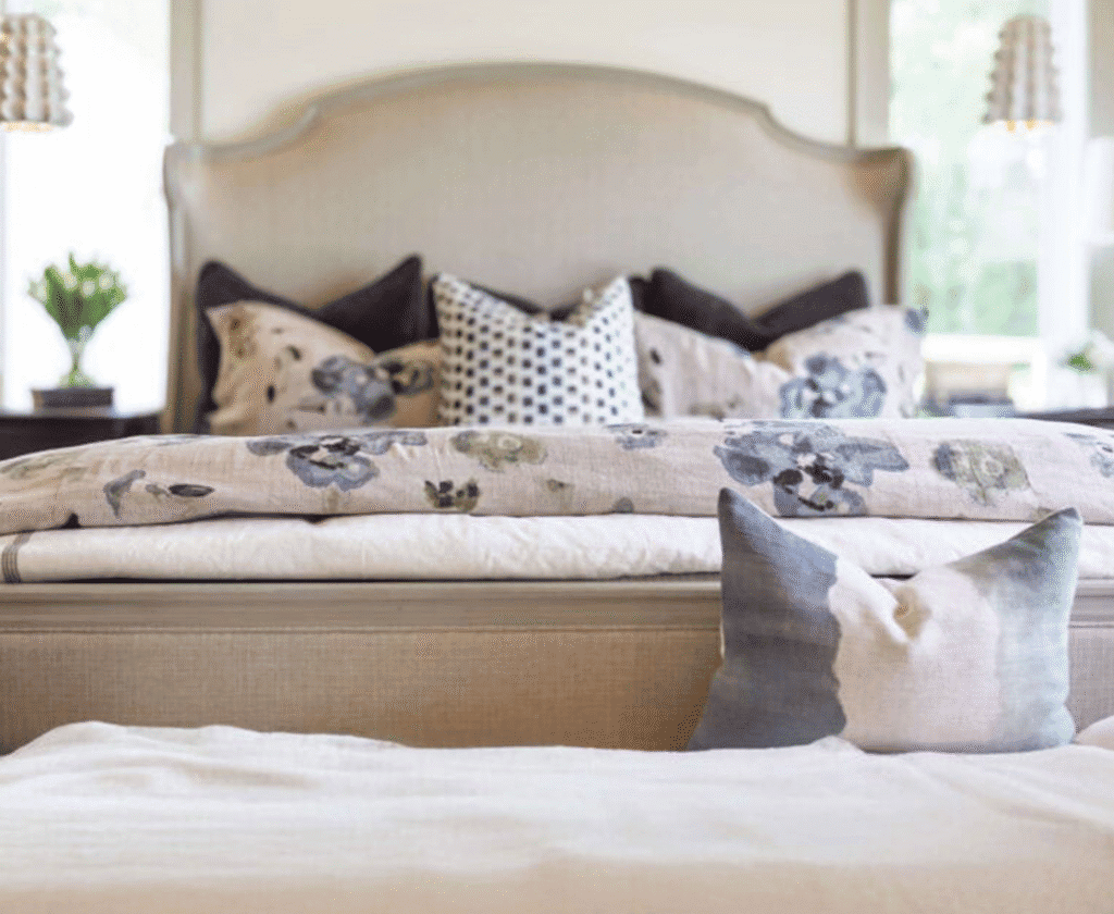 Soft Neutral-Toned Upholstered Bed With Decorative Pillows Featuring Floral And Geometric Patterns, Set In A Bright Bedroom With Large Windows And Fresh Floral Accents.
