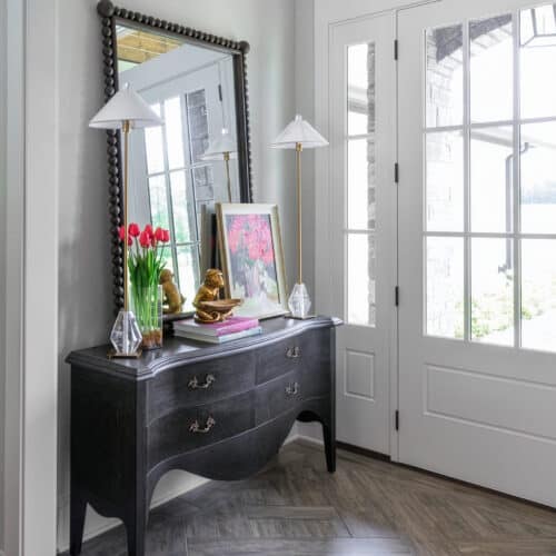 Elegant Entryway Featuring A Black Vintage-Style Console Table With Decorative Items, A Large Mirror, And Natural Light From Glass Doors, Creating A Welcoming And Stylish Home Entrance.