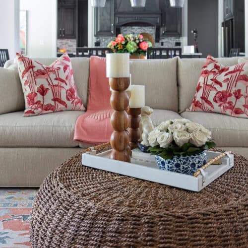 Cozy Living Room Featuring A Beige Sofa With Red Patterned Pillows, A Woven Round Coffee Table, And Decorative Candles And Flowers, Creating A Warm And Inviting Atmosphere.