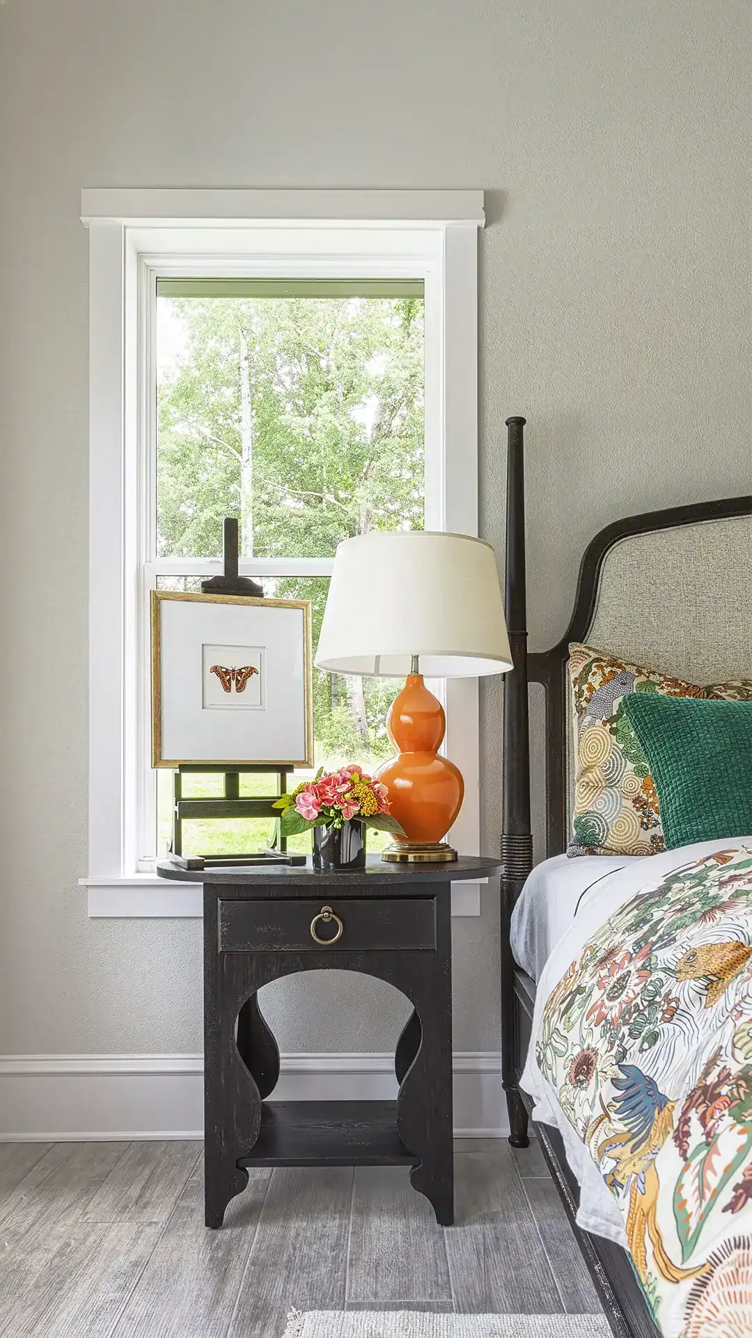 A Bright Bedroom Corner Featuring A Black Nightstand, A White Table Lamp With An Orange Base, And A Floral Bedspread. A Window With Greenery Outside Adds Natural Light To The Space.