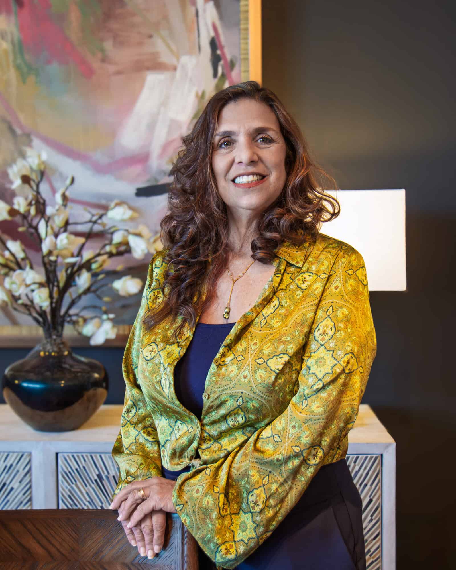 Elegant Woman With Curly Brown Hair, Wearing A Yellow Patterned Blazer And Navy Top, Standing In A Stylish Interior Setting.