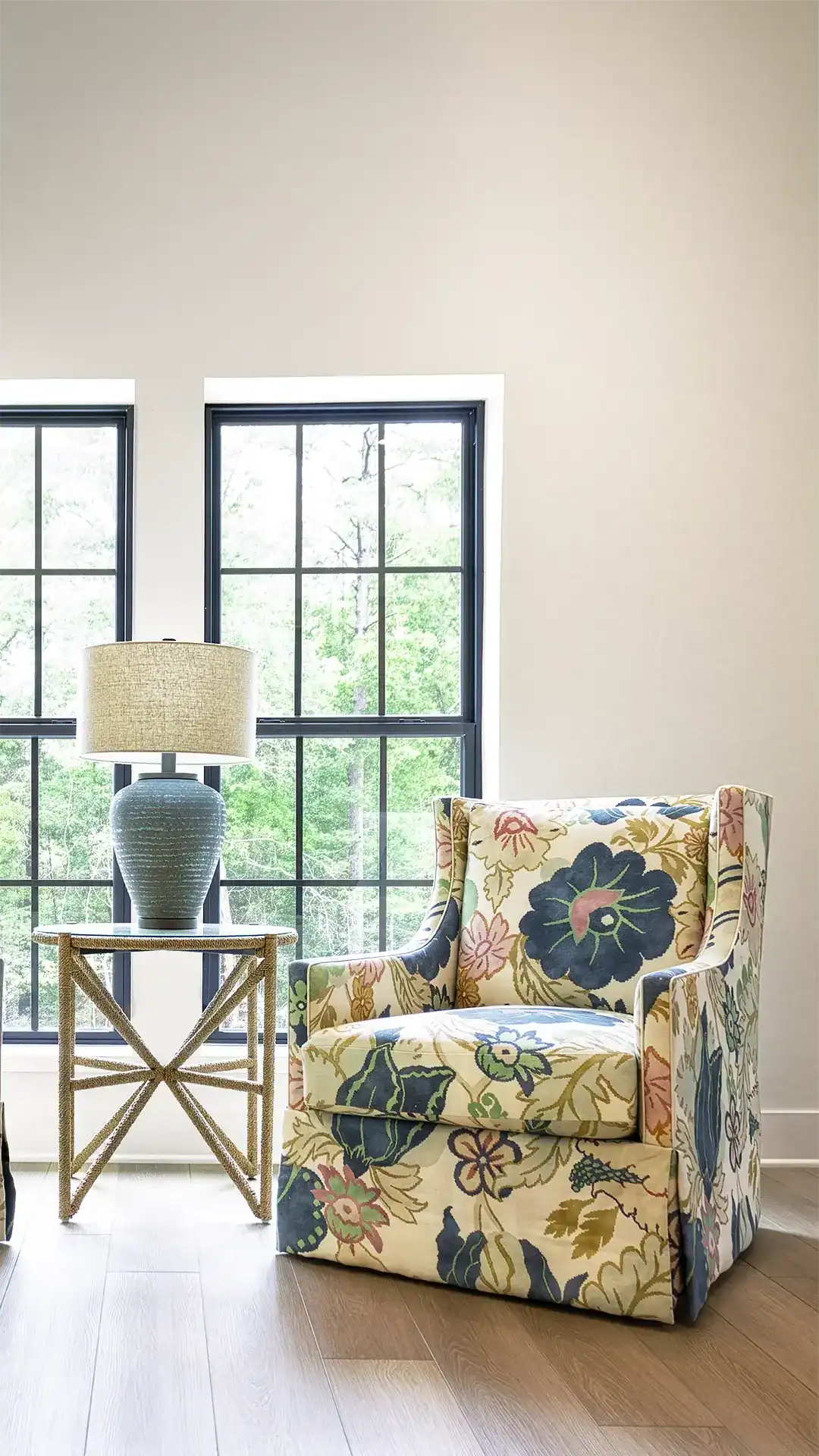A Floral Upholstered Armchair Placed Beside A Side Table With A Lamp, Set Against A Neutral Wall With Large Windows Showing Greenery Outside.