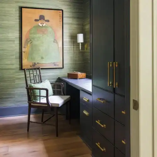Stylish Home Office Corner With Green Textured Walls, A Framed Artwork, A Wooden Desk, And A Black Cabinet With Gold Handles, Creating A Warm And Elegant Workspace.
