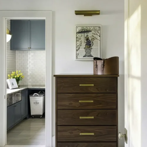 An Elegant Wooden Dresser With Gold Handles Stands Against A White Wall In A Warmly Lit Room, Complemented By Japanese-Inspired Wall Art And A Brass Wall Sconce, Creating A Serene And Inviting Atmosph.