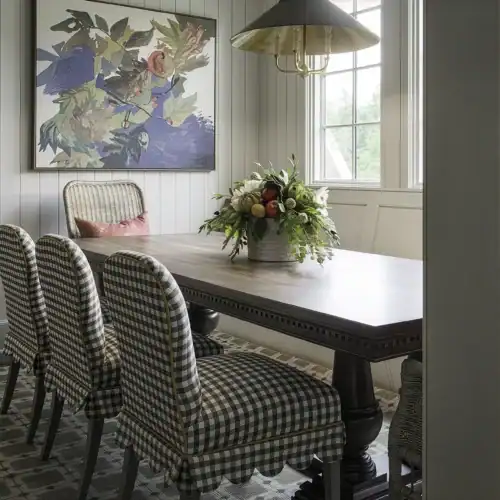 Elegant Dining Room Featuring A Dark Wood Table, Six Checkered Upholstered Chairs, A Floral Centerpiece, And A Large Colorful Abstract Painting On The Wall. Natural Light Fills The Space Through Windo.
