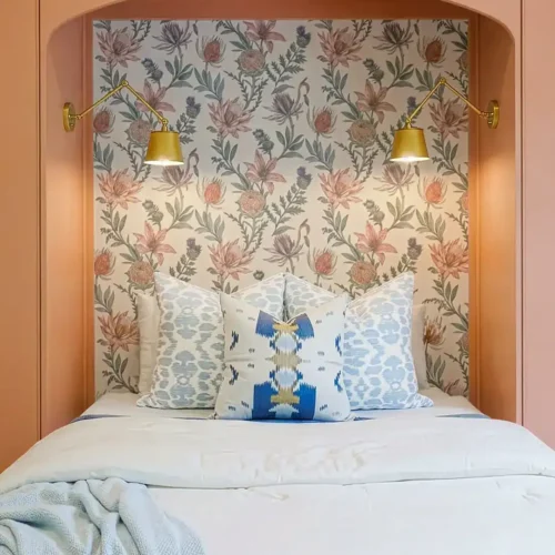 Intimate Bedroom Nook Featuring Floral Wallpaper, Two Gold Wall Sconces, And A Plush Bed With Decorative Pillows, Creating A Warm And Inviting Space.
