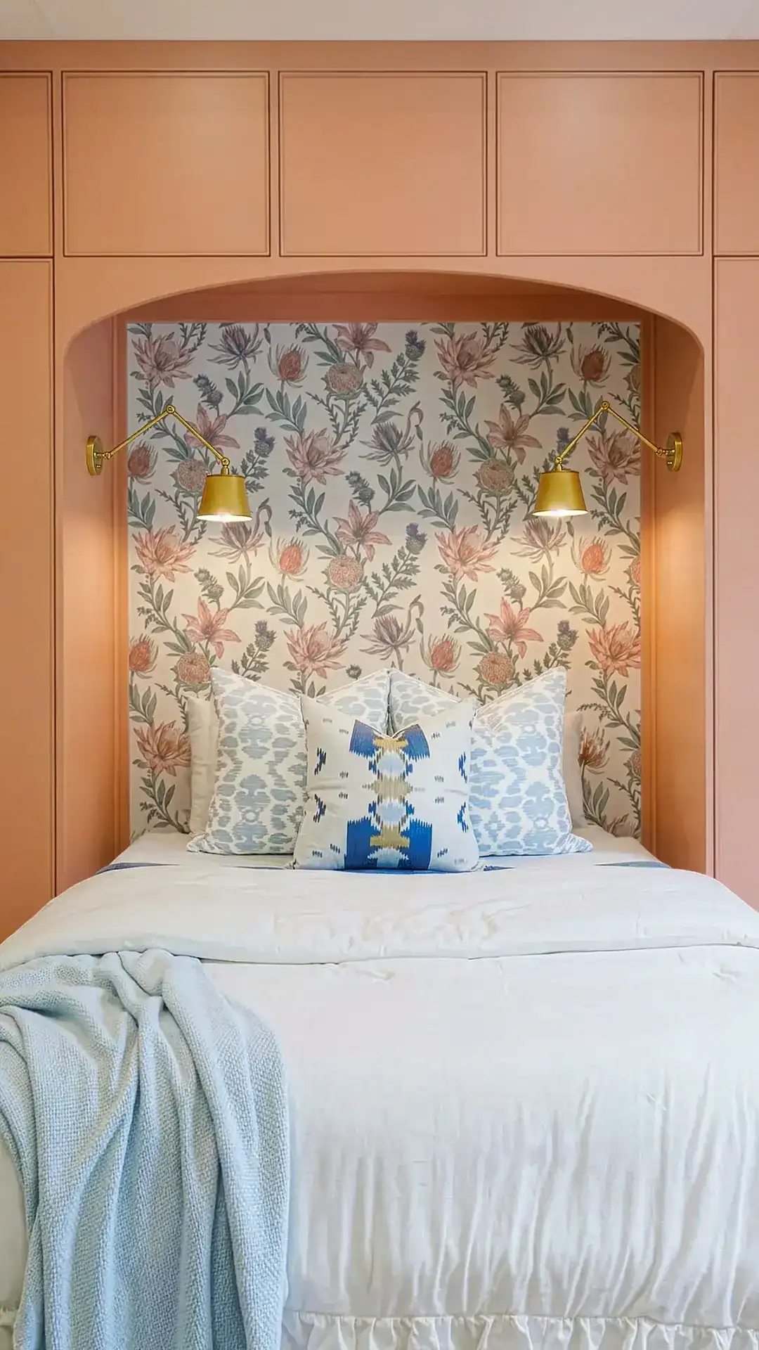 Intimate Bedroom Nook Featuring Floral Wallpaper, Two Gold Wall Sconces, And A Plush Bed With Decorative Pillows, Creating A Warm And Inviting Space.