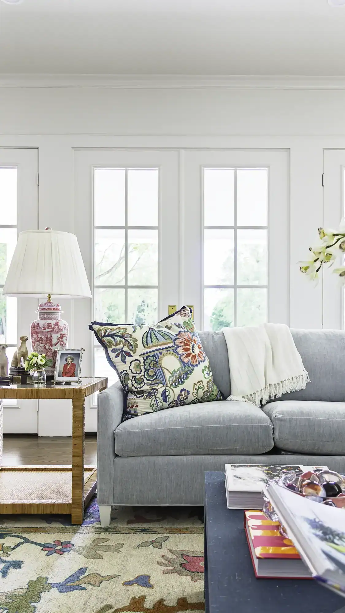 Bright Living Room Featuring A Light Blue Sofa, Floral Pillows, A White Throw Blanket, And A Side Table With A Pink Lamp And Decorative Items, Large Windows In The Background.
