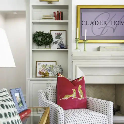 Stylish White Armchair With A Pink Decorative Pillow Featuring A Gold Dog Design, Situated In A Modern Living Room With White Shelves And A Fireplace.