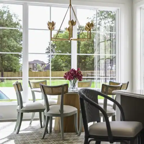 Bright Dining Room With Large Windows, Modern Chandelier, And A Round Wooden Table With Pink Flowers, Surrounded By Mixed Chairs.