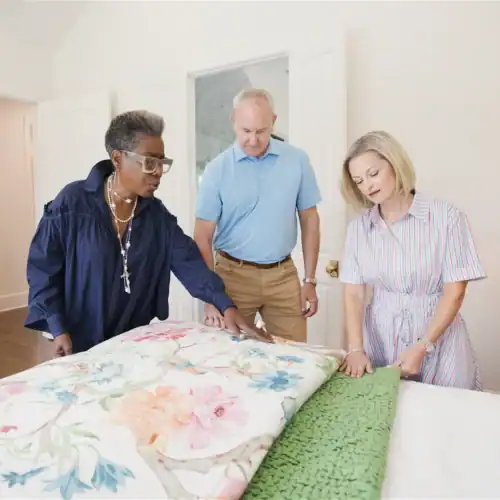 Three People Examining A Floral Bedspread In A Furniture Store, Showcasing Home Decor Options In Tyler, Texas.